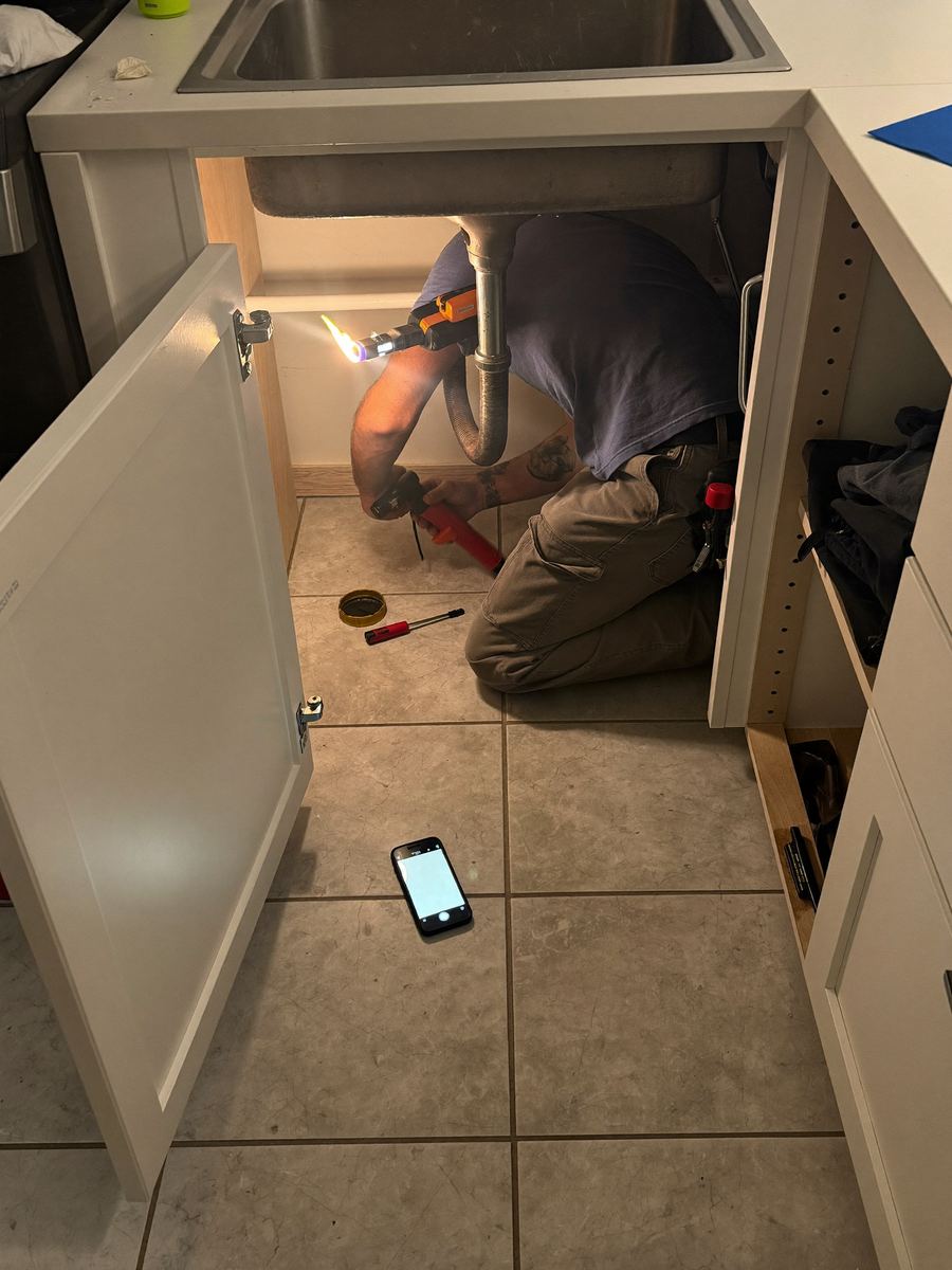 A plumber under a kitchen sink, phone lit up on the floor beside him